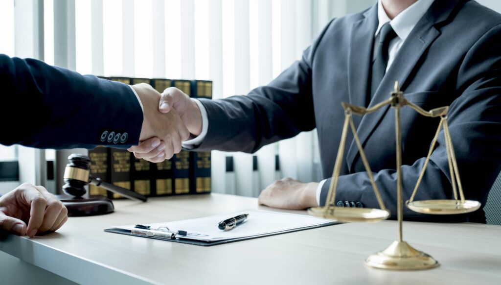 Personal injury attorney shaking hands with client in law office, with gavel and scales of justice on desk.