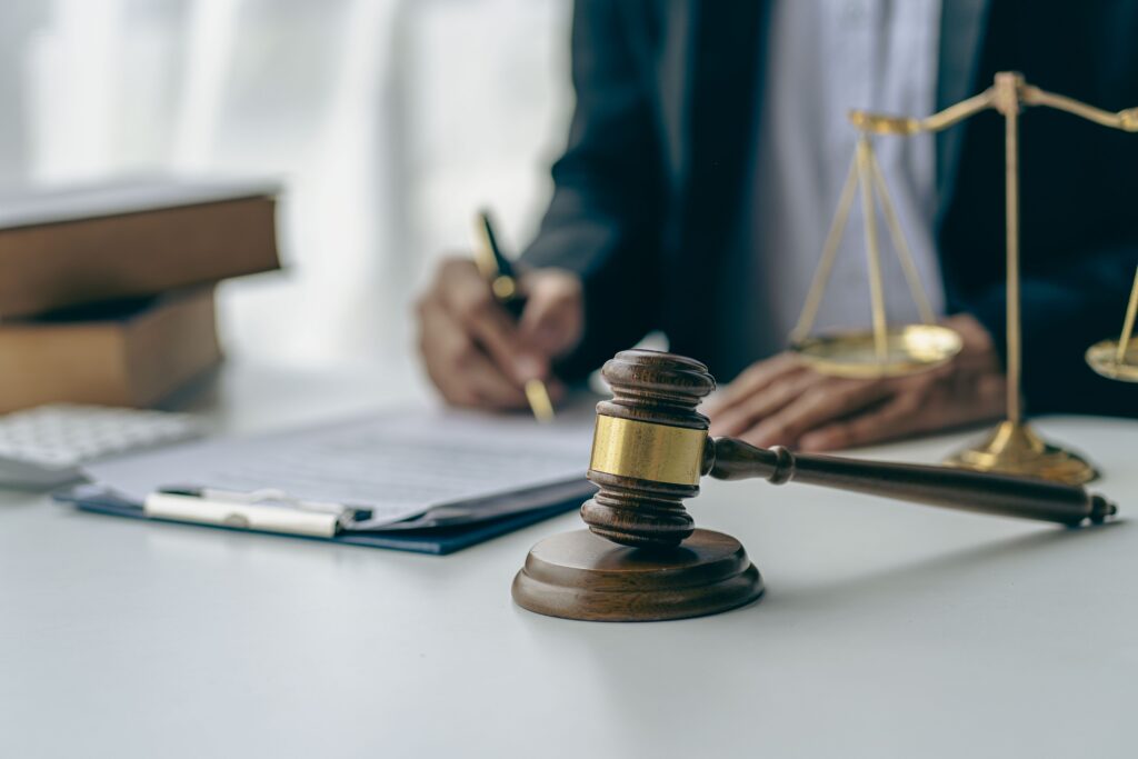 Judge&rsquo;s gavel and scales of justice on desk with lawyer signing legal documents in background, symbolizing courtroom proceedings and legal services.