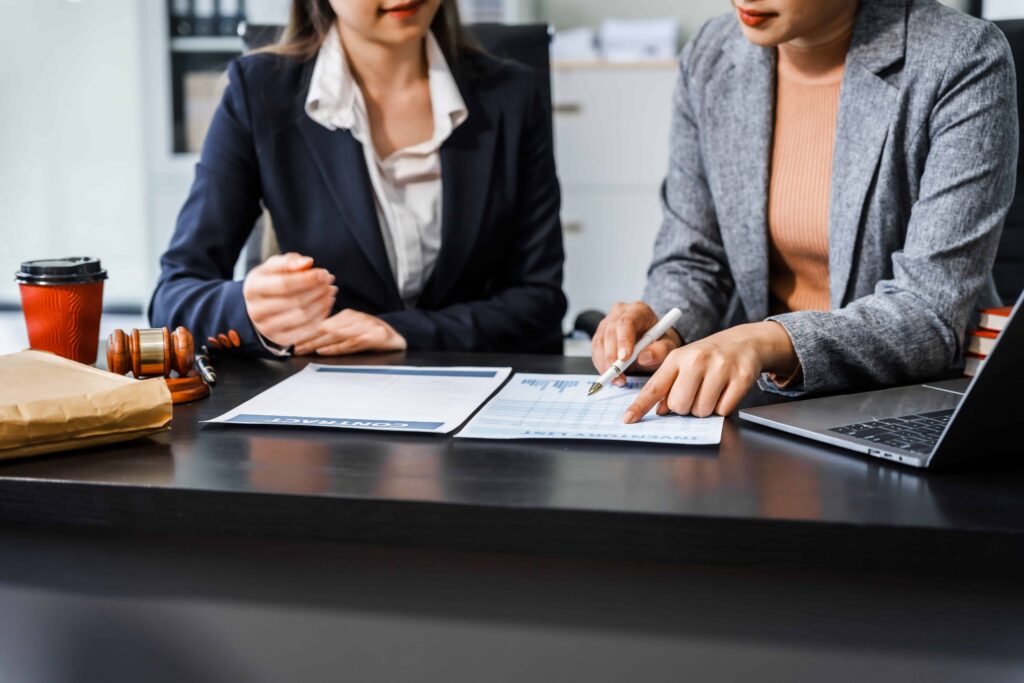 Two female lawyers reviewing contract documents at office desk with gavel and laptop, discussing legal agreement details.