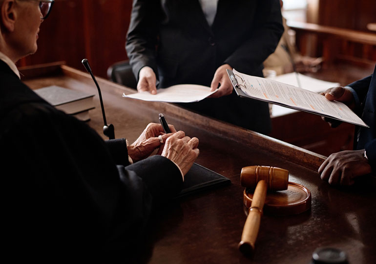 A judge examines documents at the courtroom bench while attorneys present case files during a business litigation proceeding, with a gavel placed on the desk.