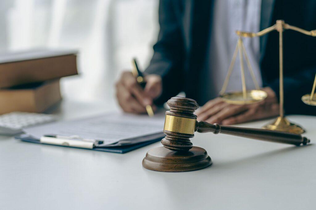 Judge&rsquo;s gavel and scales of justice on desk with lawyer signing legal documents in background, symbolizing courtroom proceedings and legal services.
