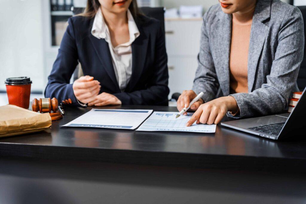 Two female lawyers reviewing contract documents at office desk with gavel and laptop, discussing legal agreement details.