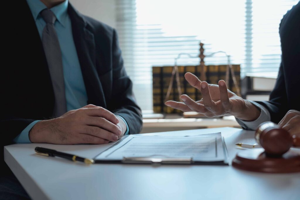 Lawyer discussing legal case with client at office desk, with contract paperwork, pen, and judge&rsquo;s gavel in foreground.