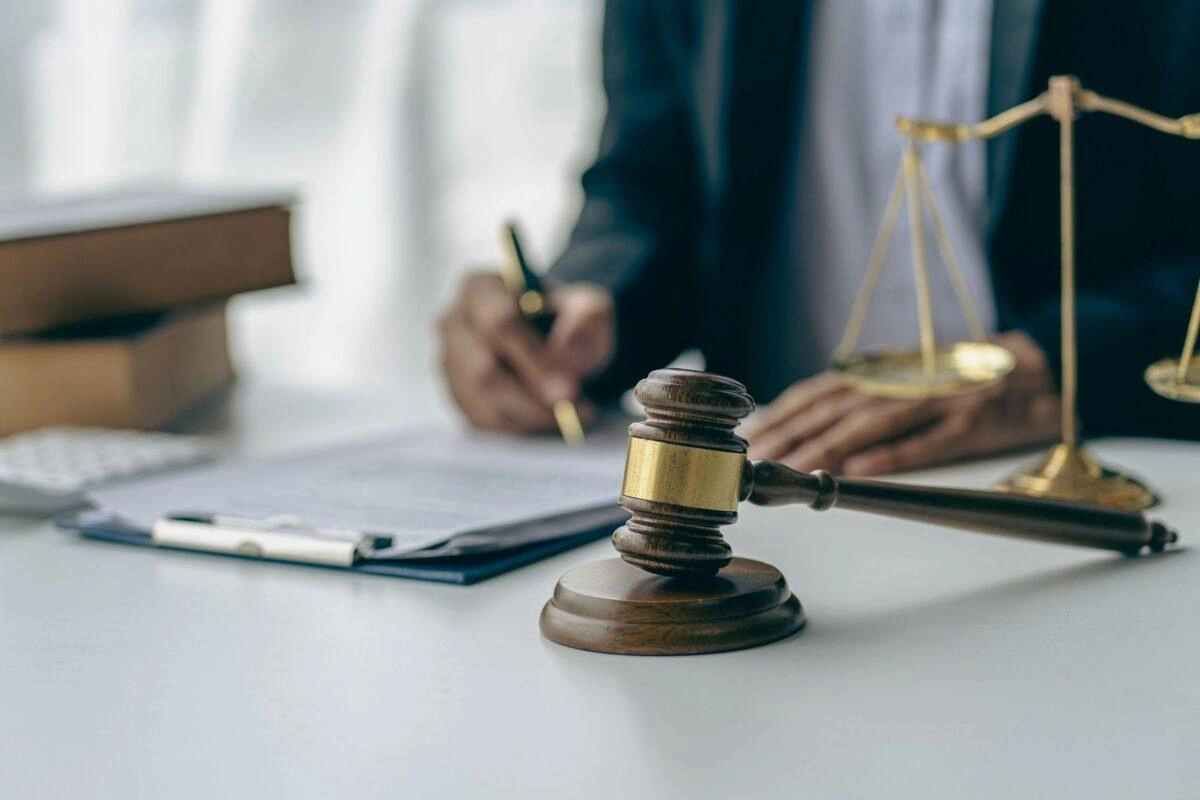 Judge’s gavel and scales of justice on desk with lawyer signing legal documents in background, symbolizing courtroom proceedings and legal services.