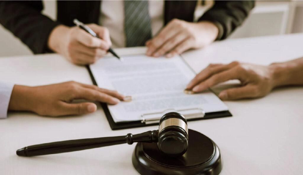 Clients signing legal agreement with attorney at desk, judge’s gavel in foreground representing settlement or lawsuit resolution.